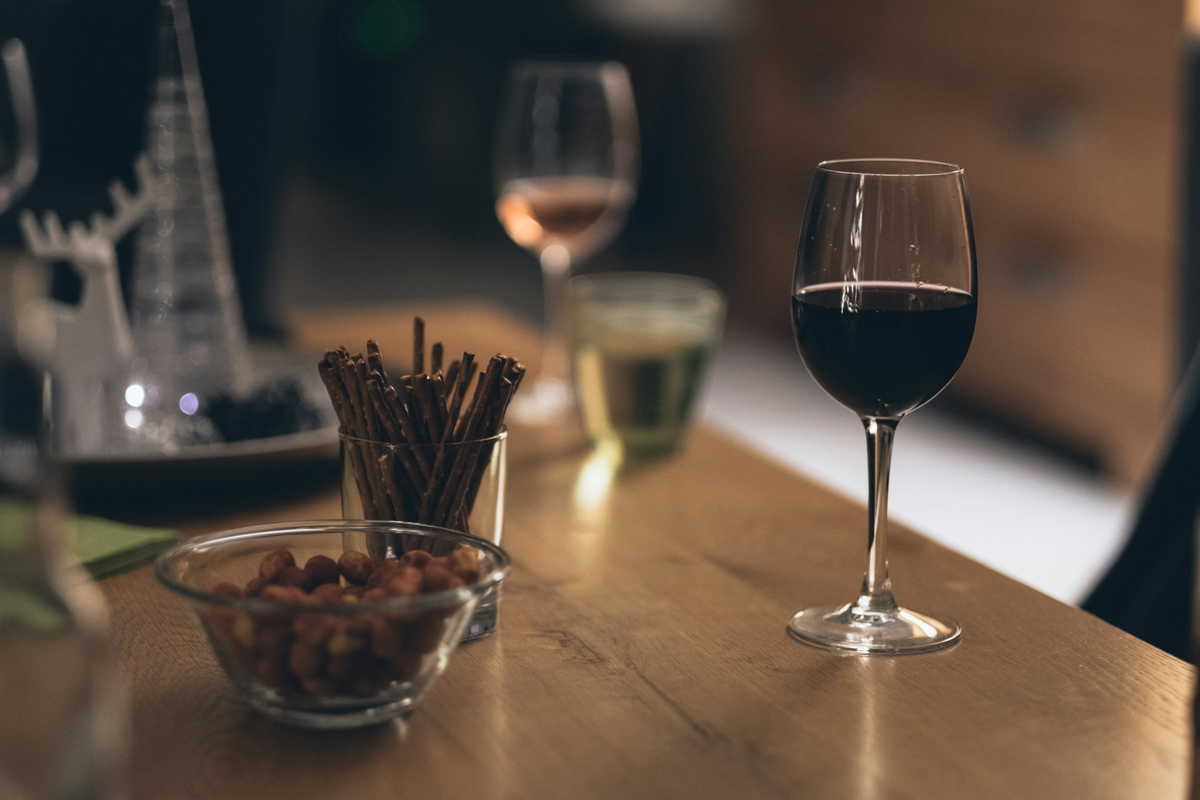 Taça de vinho tinto em primeiro plano, com petiscos e palitos salgados sobre mesa de madeira.