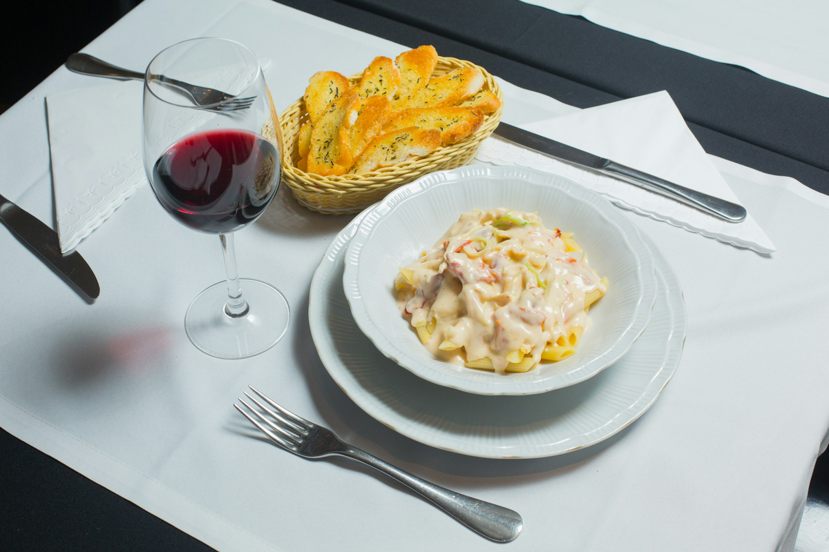Penne com molho cremoso e vegetais, torradas e taça de vinho tinto em mesa posta.