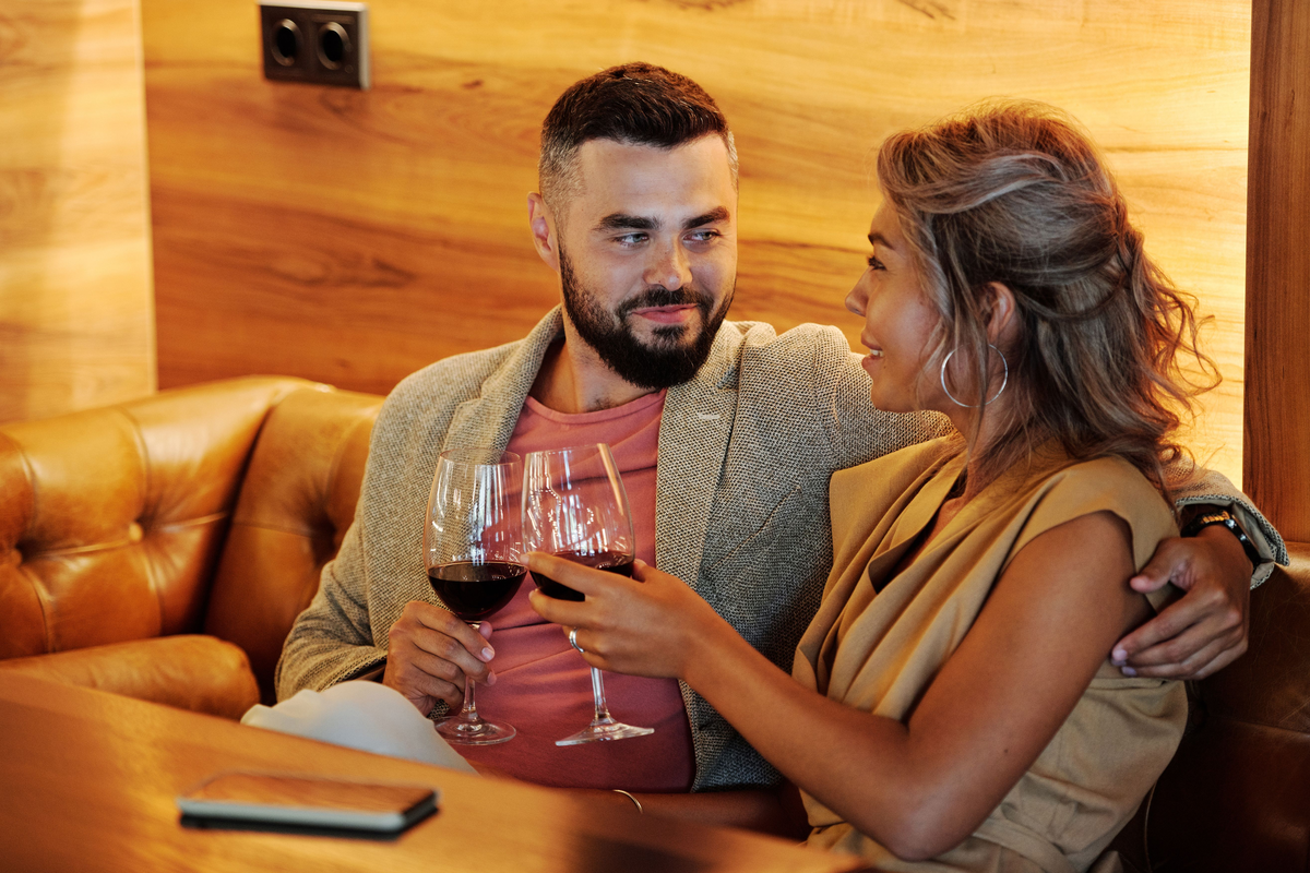 Casal sentado junto em um restaurante, segurando taças de vinho tinto diante de uma mesa de madeira.