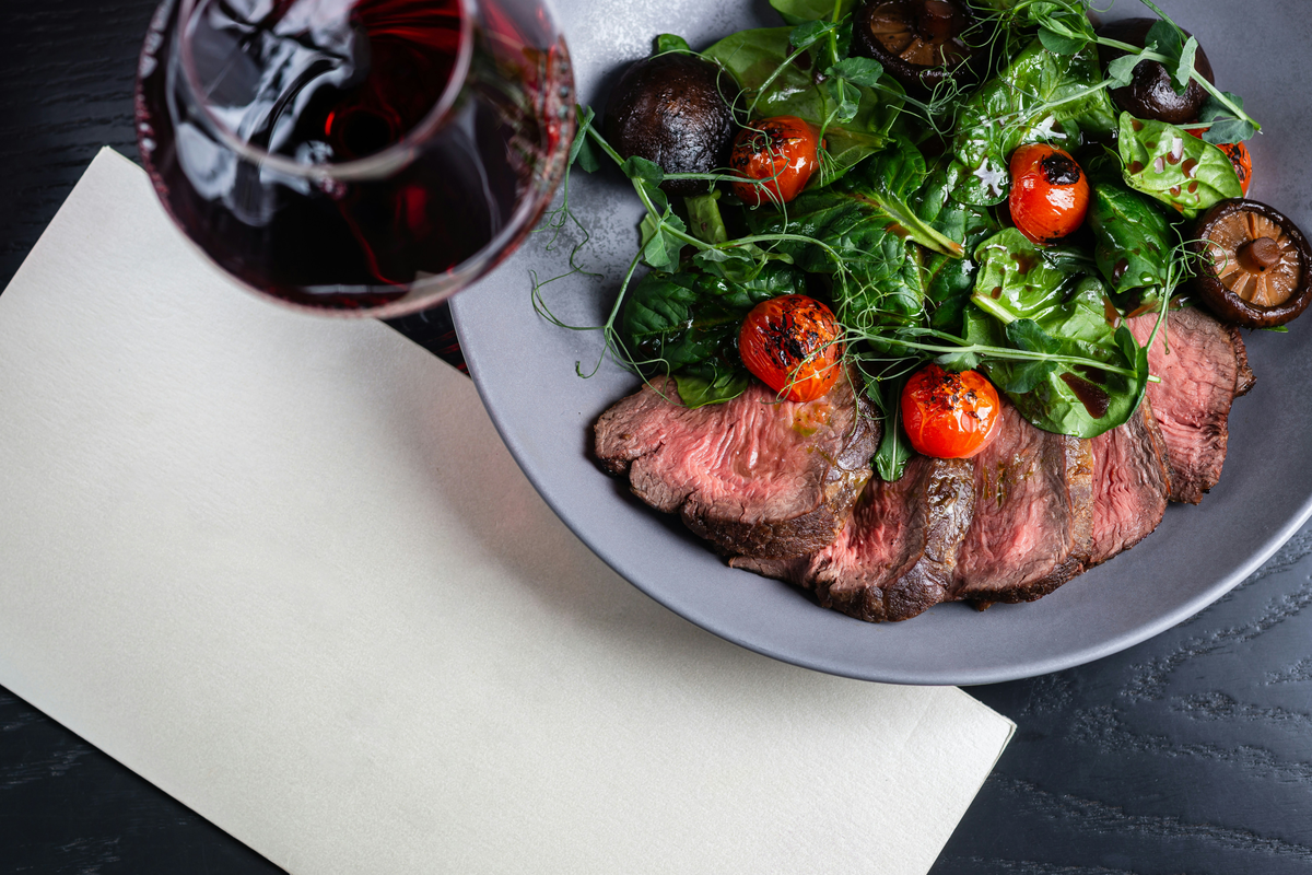 Fatias de carne bovina com folhas, tomates e cogumelos; taça de vinho tinto parcialmente enquadrada no canto superior.