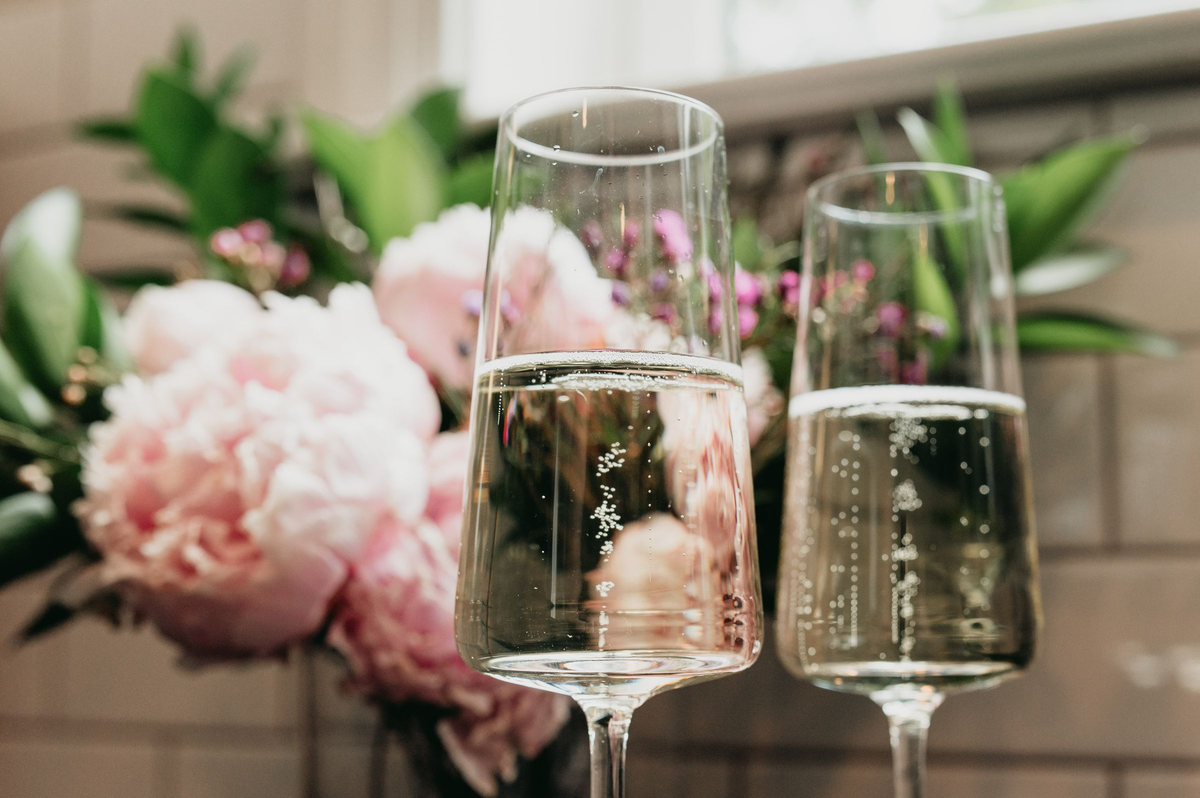 Duas taças com espumante claro e bolhas, ao lado de um arranjo de flores claras.