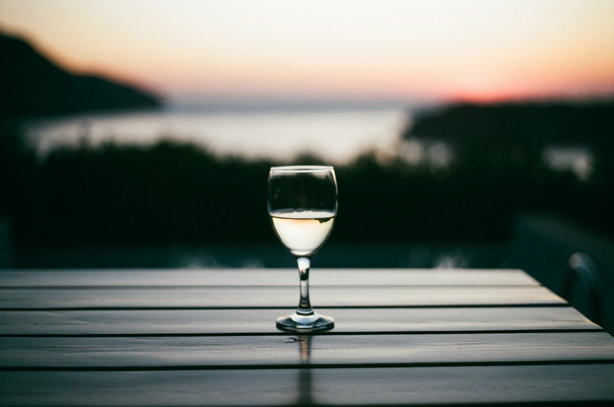 Taça de vinho branco claro em mesa de madeira com litoral ao fundo.