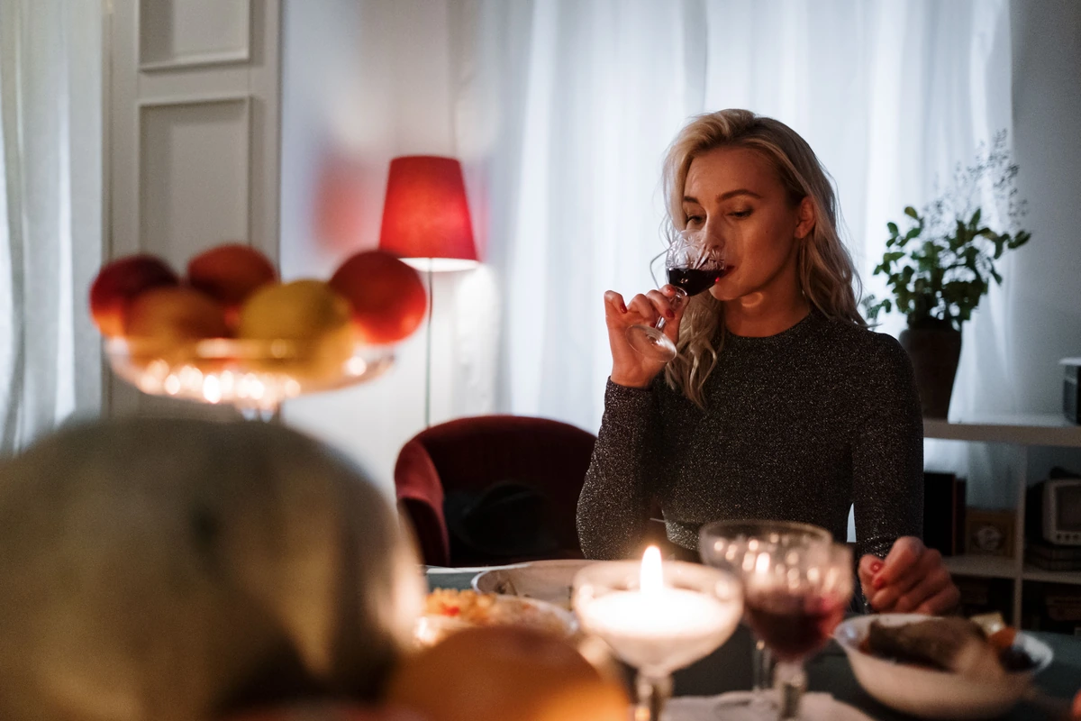 Mulher adulta aprecia vinho tinto à mesa com vela acesa, pães, frutas e pratos.