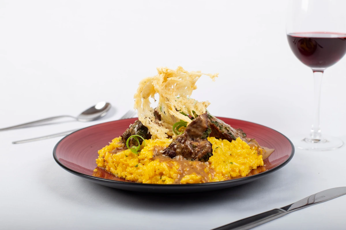 Prato vermelho com carne ao molho, risoto e crocante de queijo; taça de vinho tinto ao fundo.