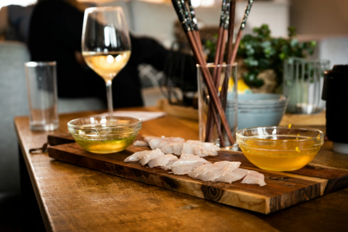 Peixe cru fatiado em tábua, molhos, hashi e taça de vinho branco.