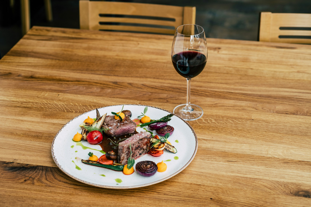 Carne com legumes coloridos e taça de vinho tinto sobre mesa de madeira.