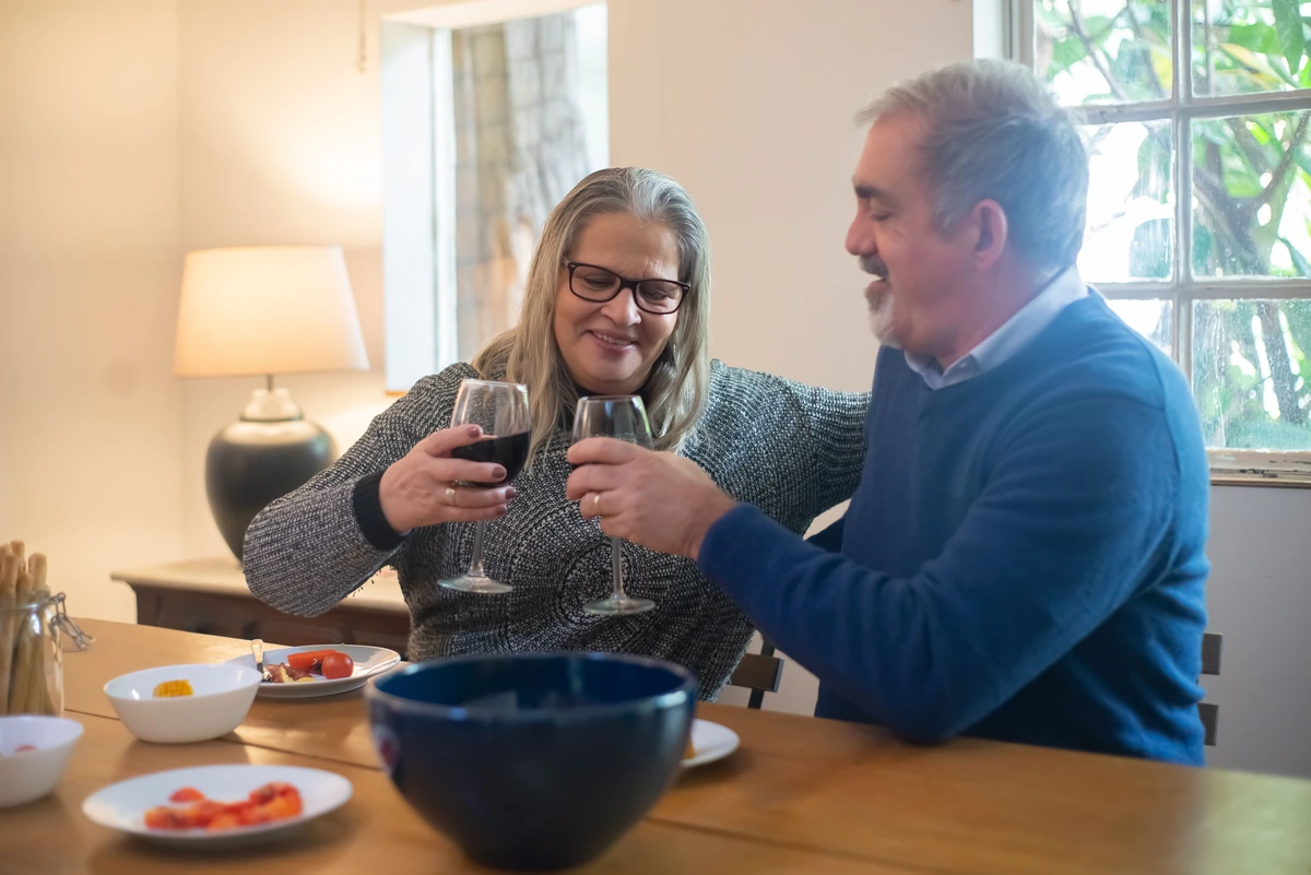 Casal à mesa aproximando duas taças preenchidas de vinho tinto.