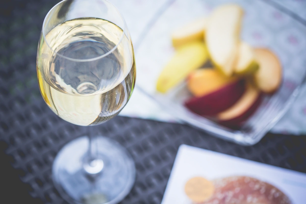 Taça de vinho branco claro, com frutas e aperitivos desfocados sobre a mesa.