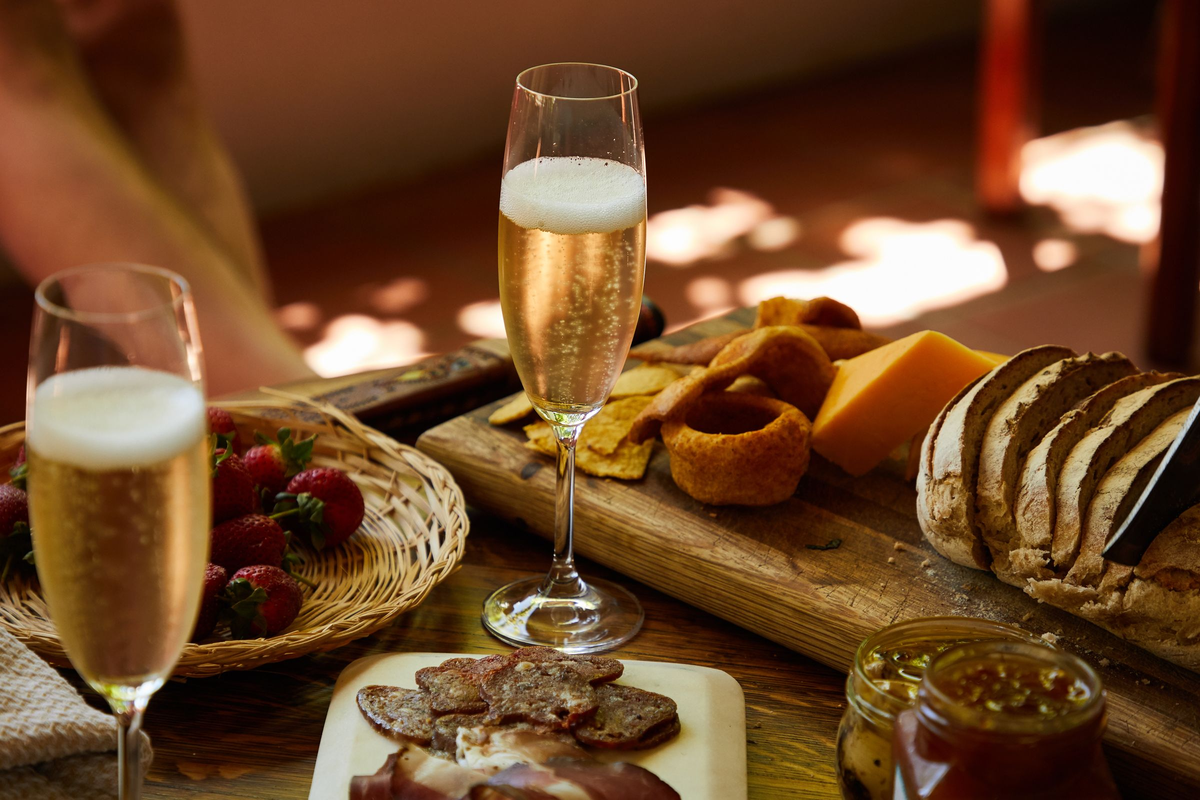 Duas flutes de espumante junto a queijo, morangos, pães e aperitivos sobre mesa de madeira.