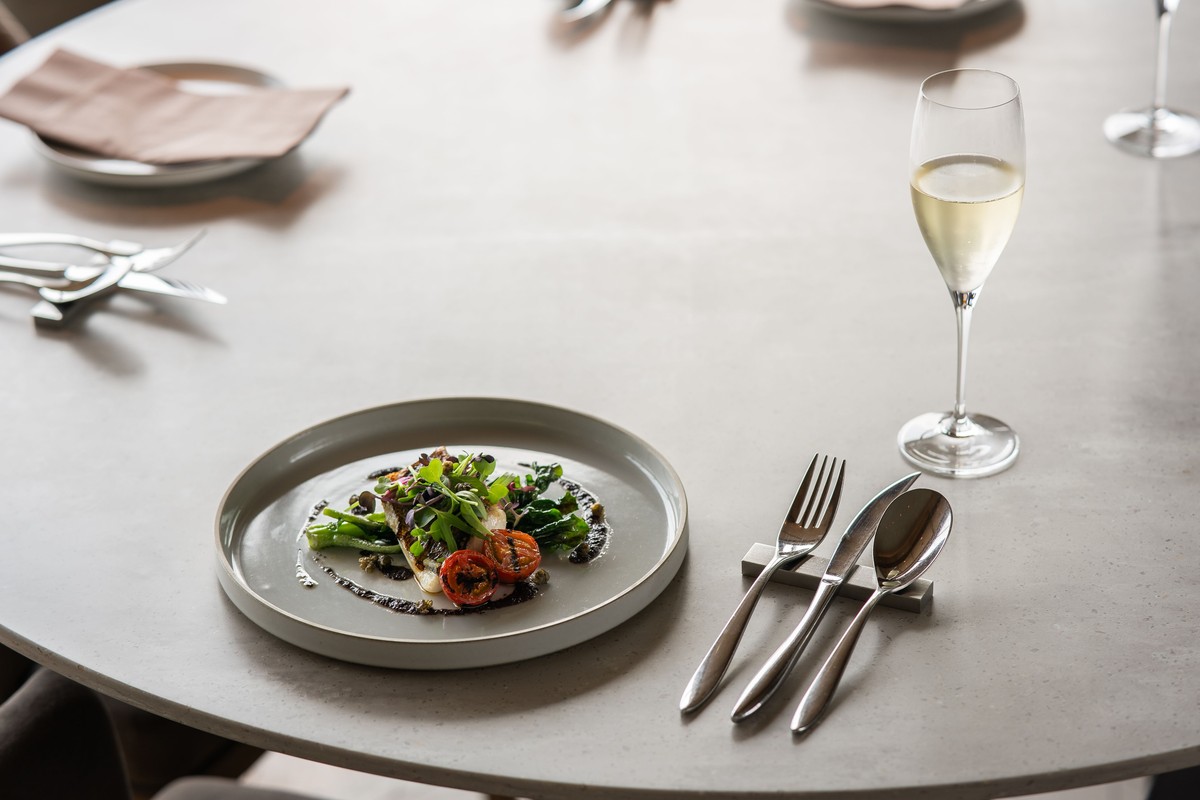 Peixe com folhas verdes e tomates ao lado de uma flûte preenchida de espumante branco.