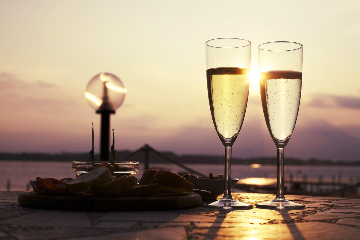Duas taças de espumante branco com aperitivos sobre mesa ao pôr do sol.