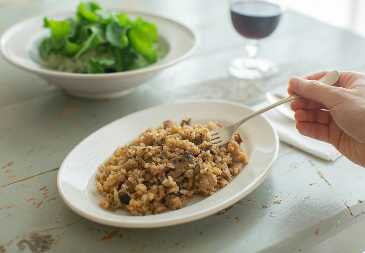 Prato de arroz com cogumelos, salada ao fundo e taça com vinho tinto sobre mesa clara.