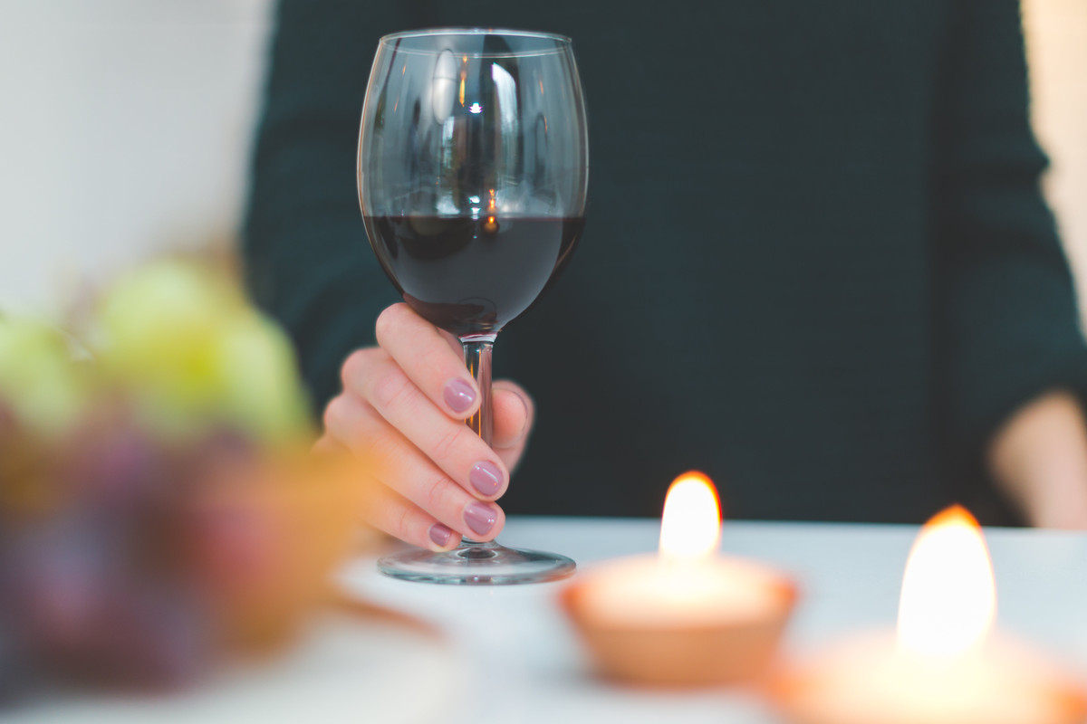 Pessoa segurando uma taça de vinho tinto, com velas acesas e frutas desfocadas sobre a mesa.