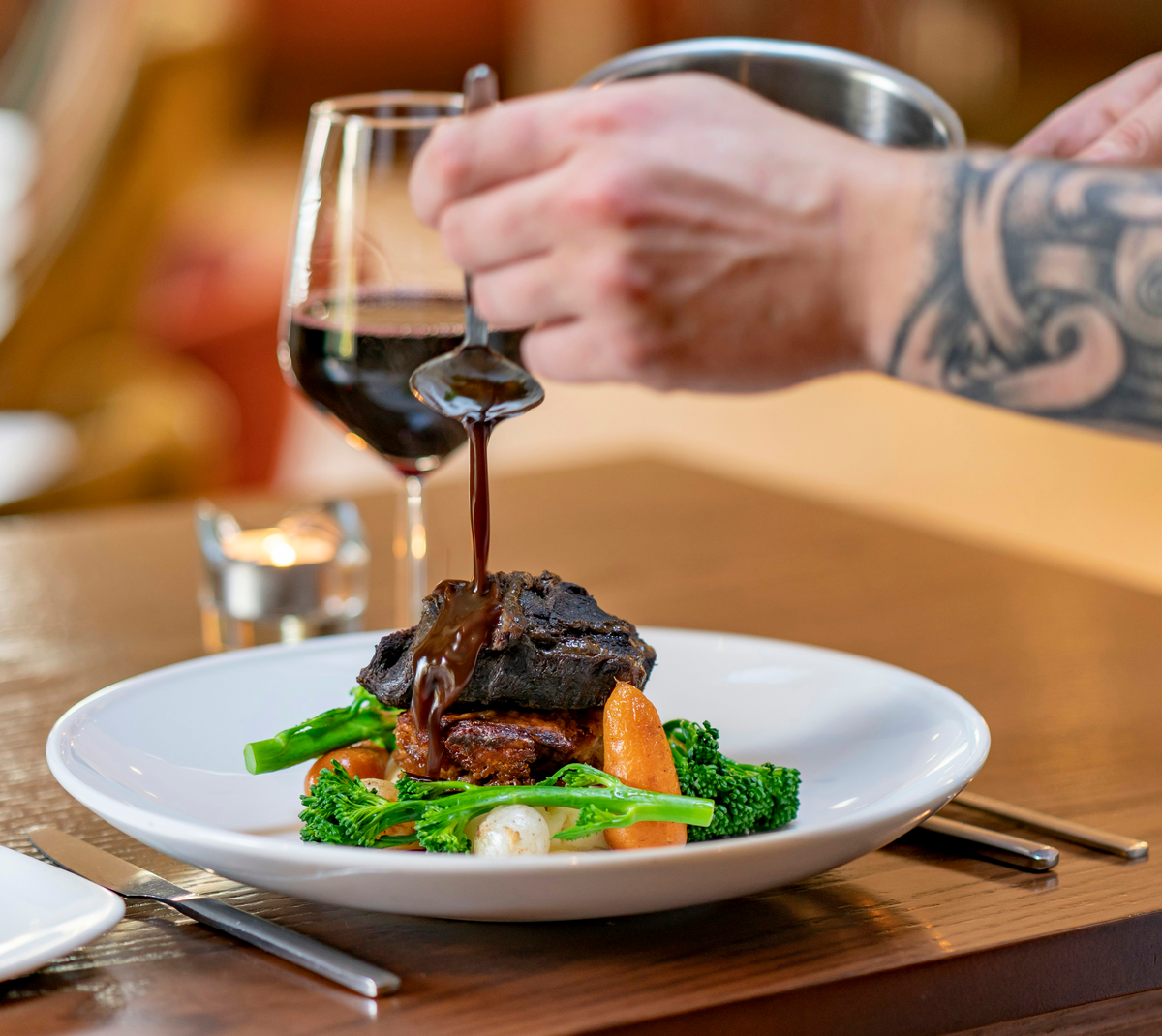 Mão acrescentando molho à carne com cenouras e vegetais; taça de vinho tinto parcialmente visível ao fundo.