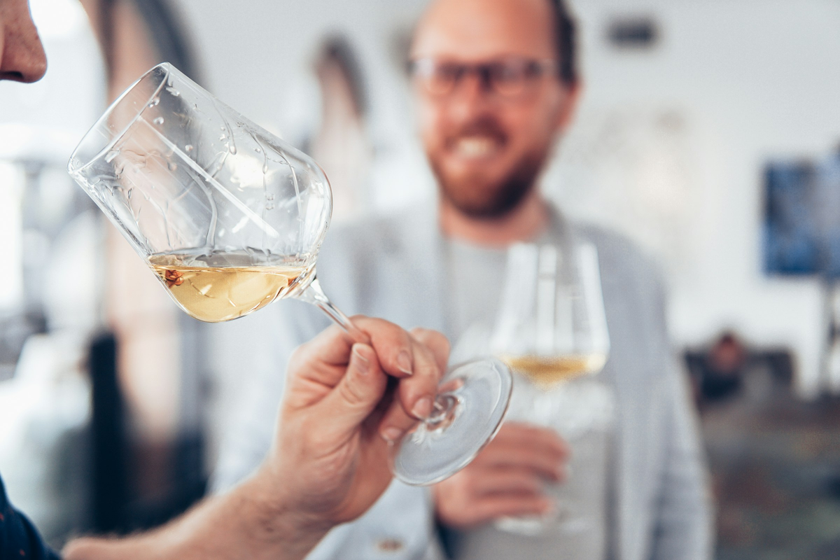 Pessoas adultas em uma degustação de vinho branco, com taça em primeiro plano.