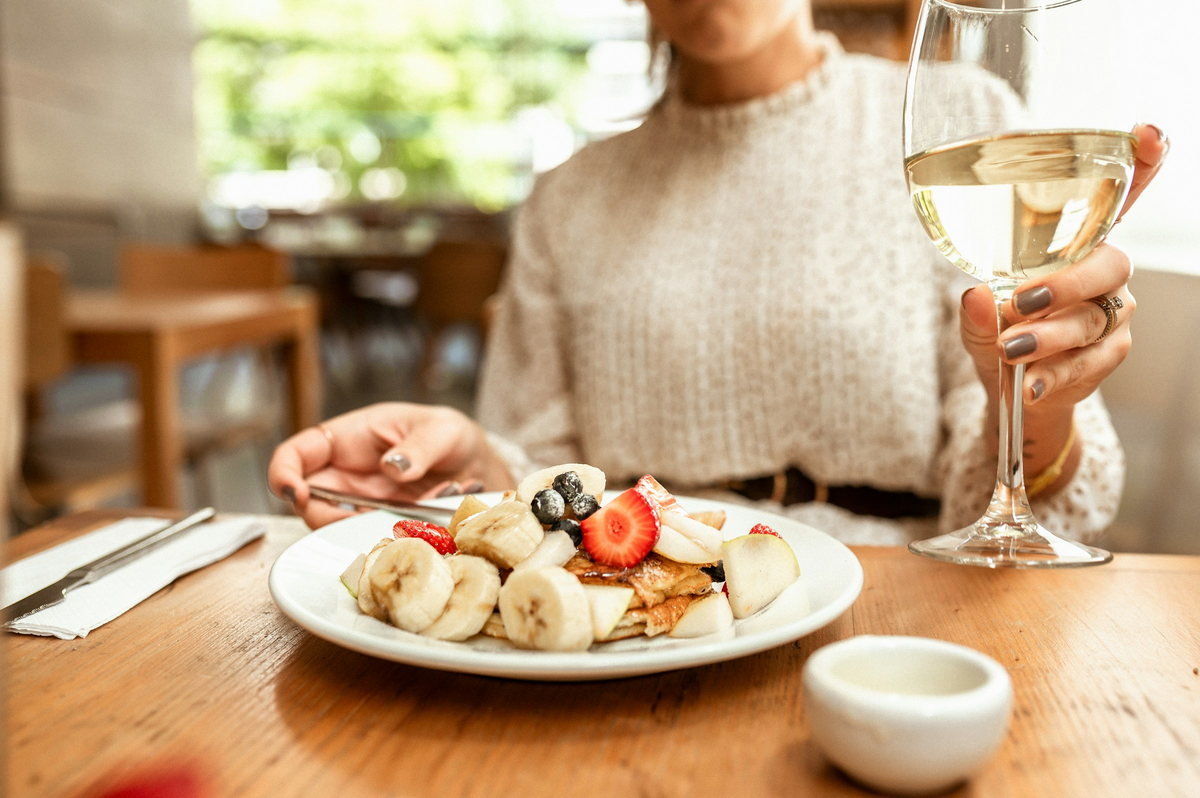 Mesa com panquecas, banana, morangos e outras frutas, com pessoa segurando taça de vinho branco.