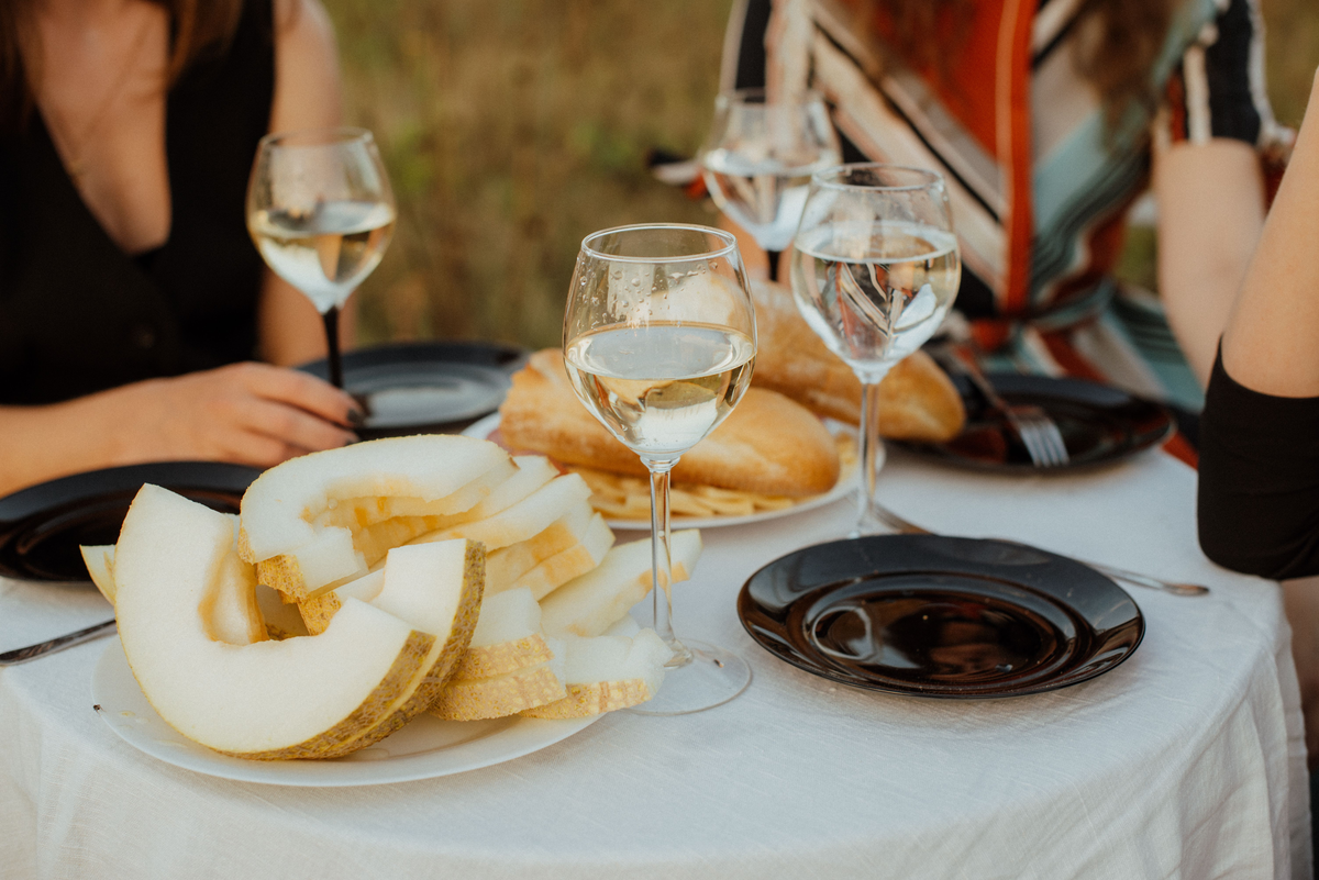 Pessoas à mesa externa com vinho branco, melão fatiado e pão sobre toalha branca.