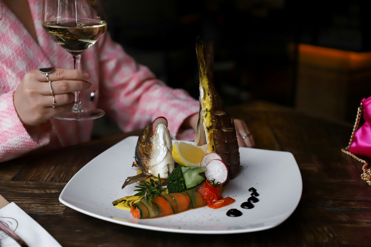 Pessoa segurando vinho branco junto a um prato de peixe com vegetais, limão e molho decorativo.