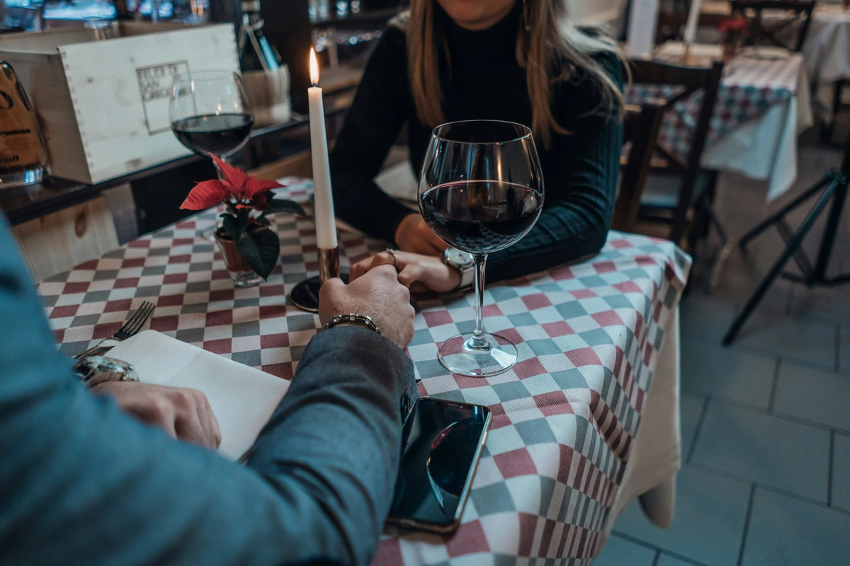 Casal de mãos dadas à mesa, com taças de vinho tinto e vela acesa.