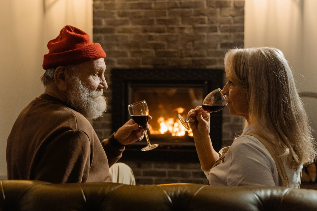 Casal adulto diante de uma lareira com taças preenchidas de vinho tinto; a mulher leva a taça aos lábios.