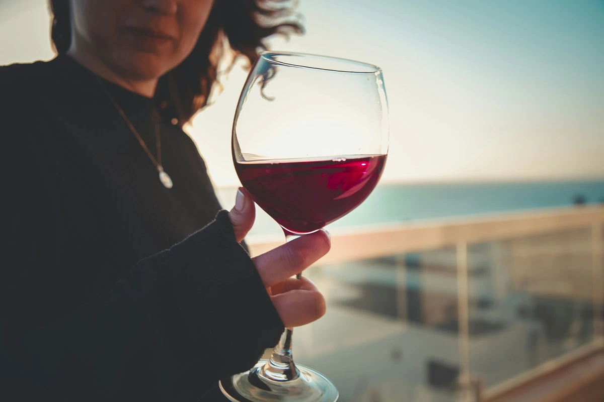 Mulher adulta segura uma taça de vinho tinto rubi translúcido ao ar livre, com céu e água desfocados ao fundo.
