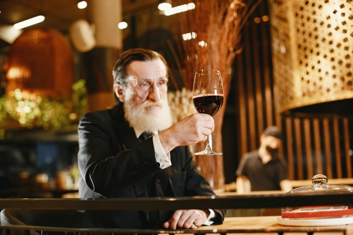 Homem adulto observa uma taça de vinho tinto em um restaurante com madeira e iluminação quente.