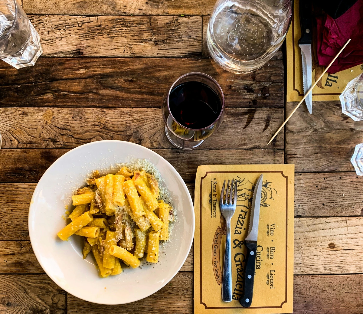Massa à carbonara em prato branco, taça de vinho tinto e copos de água em uma mesa de madeira.