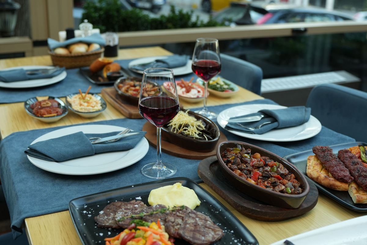Mesa com carnes grelhadas, legumes e duas taças de vinho tinto.