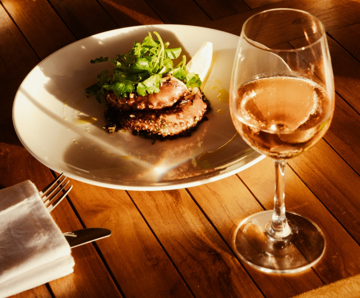 Polvo grelhado e salada em prato, com taça preenchida de vinho rosé na mesma mesa.