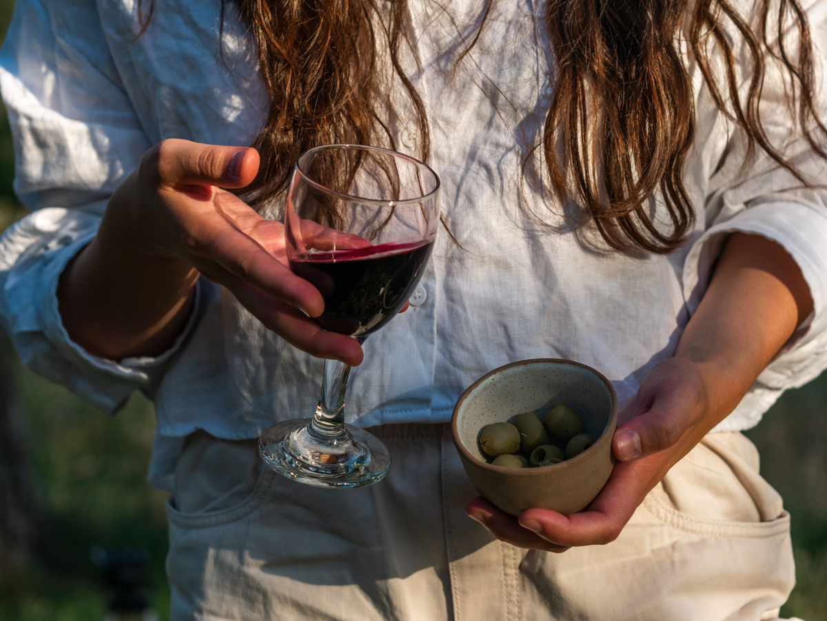 Pessoa adulta segura uma taça de vinho tinto e uma pequena tigela de azeitonas verdes.