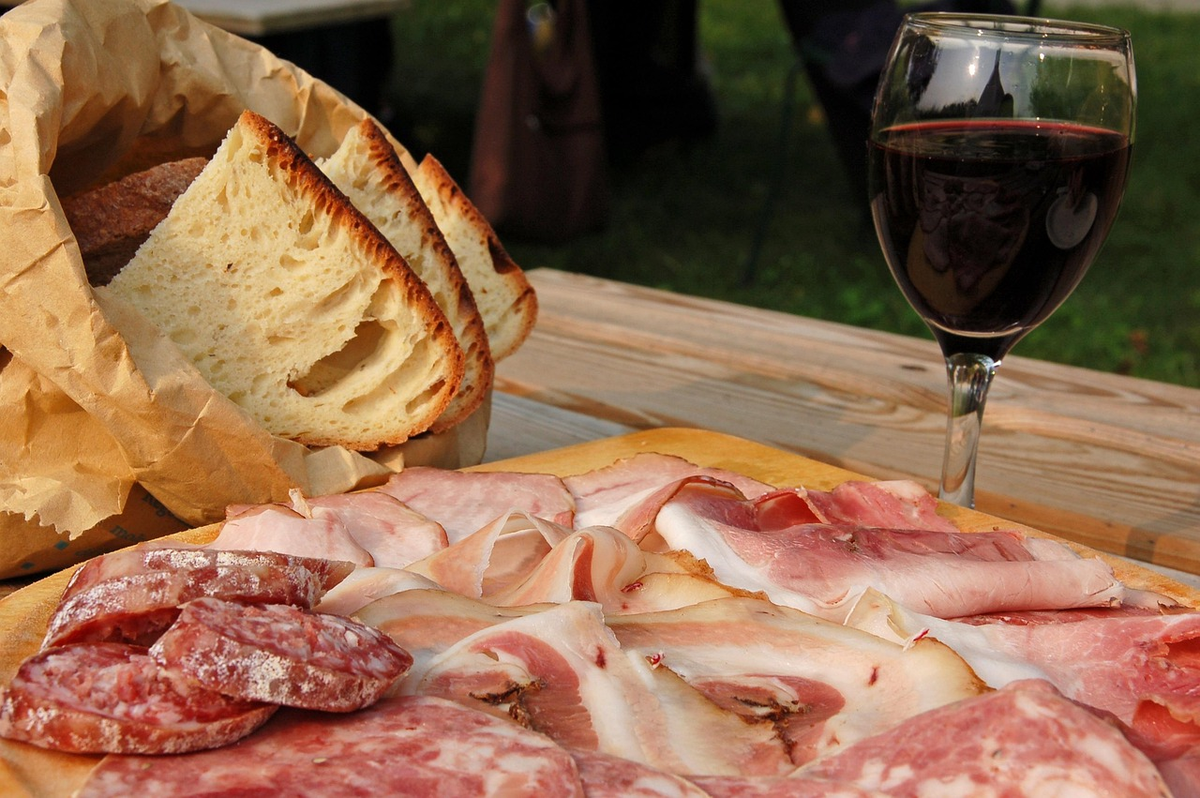 Tábua de embutidos fatiados, pão e uma taça de vinho tinto ao ar livre.