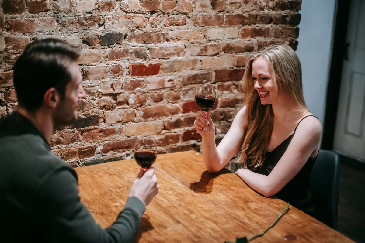 Casal conversando à mesa, cada pessoa com uma taça de vinho tinto.