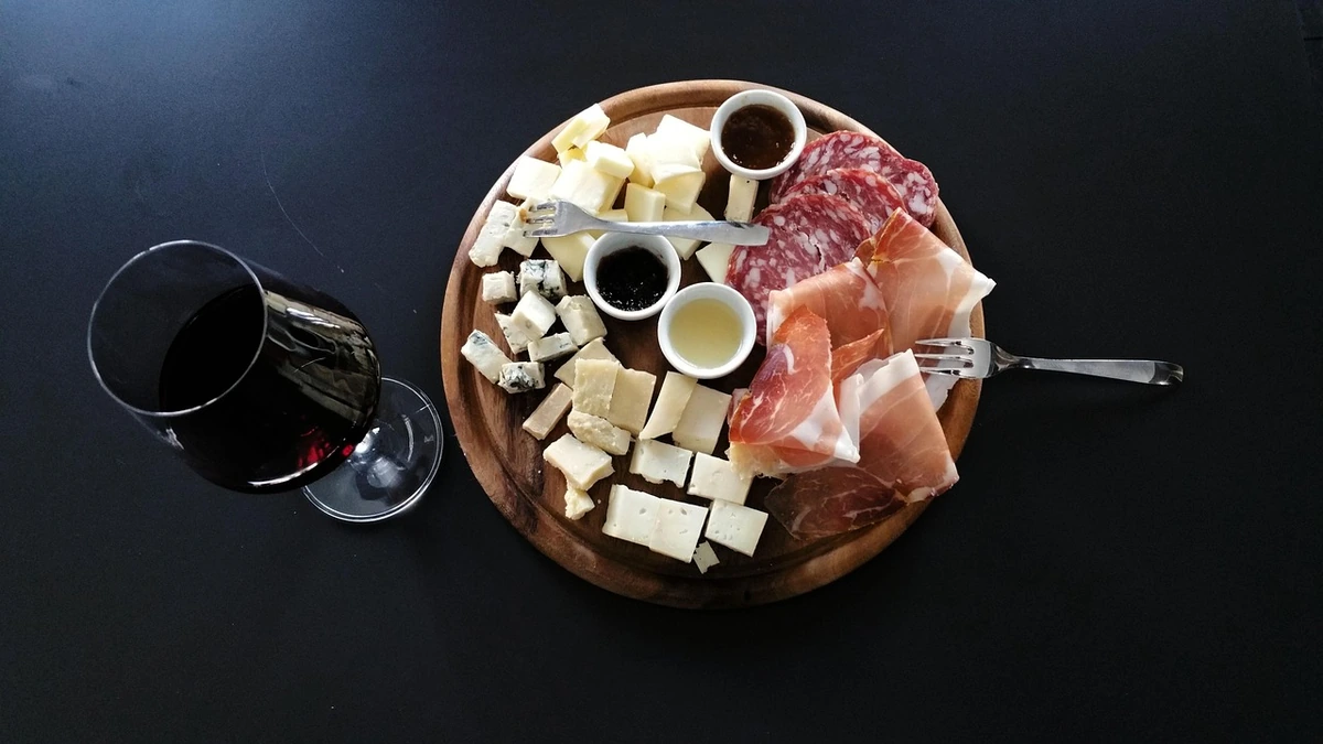 Tábua oval com queijos, salame, presunto e acompanhamentos, ao lado de uma taça de vinho tinto.