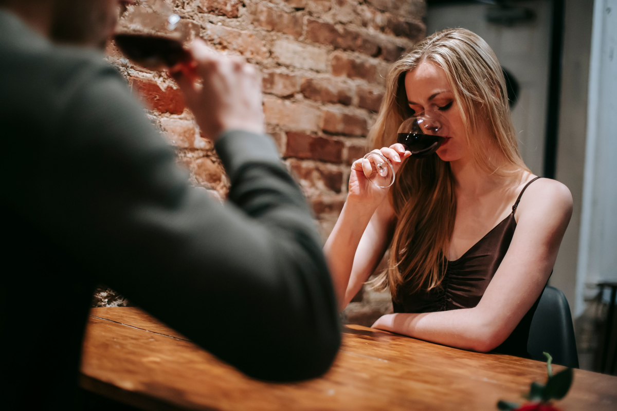 Mulher de olhos fechados provando vinho tinto à mesa, com outra pessoa e uma taça desfocadas em primeiro plano.