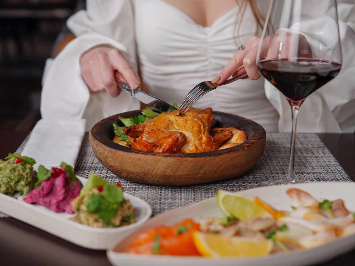 Pessoa cortando frango assado servido em recipiente de madeira, com uma taça grande de vinho tinto à direita.