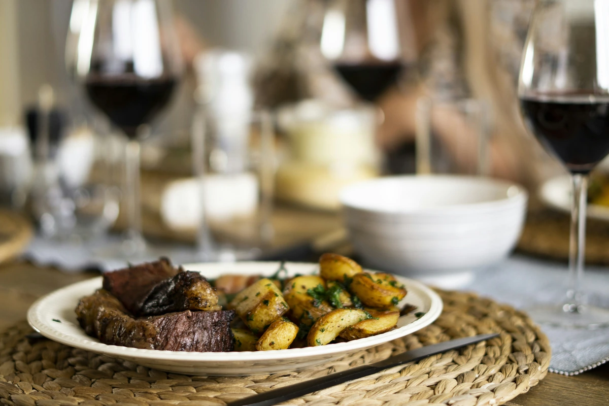 Carne bovina e batatas no prato, com taças de vinho tinto ao fundo.