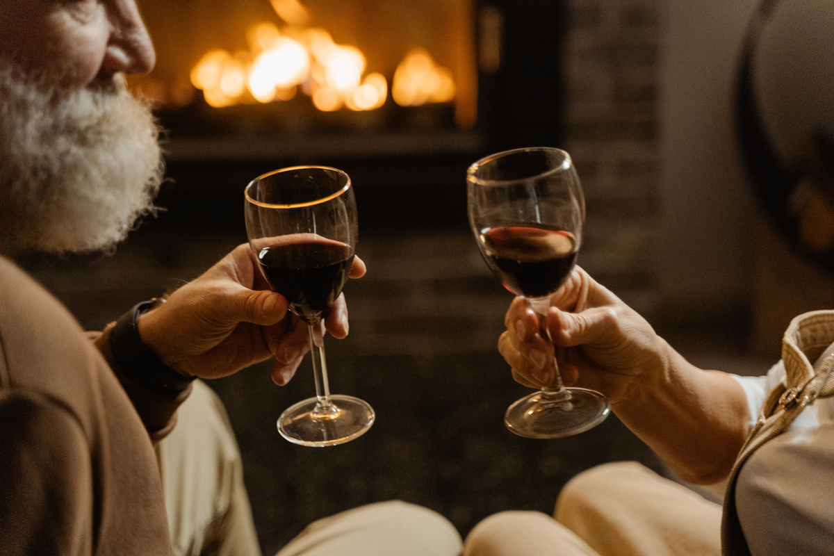 Duas pessoas seguram taças de vinho tinto diante de uma lareira desfocada.
