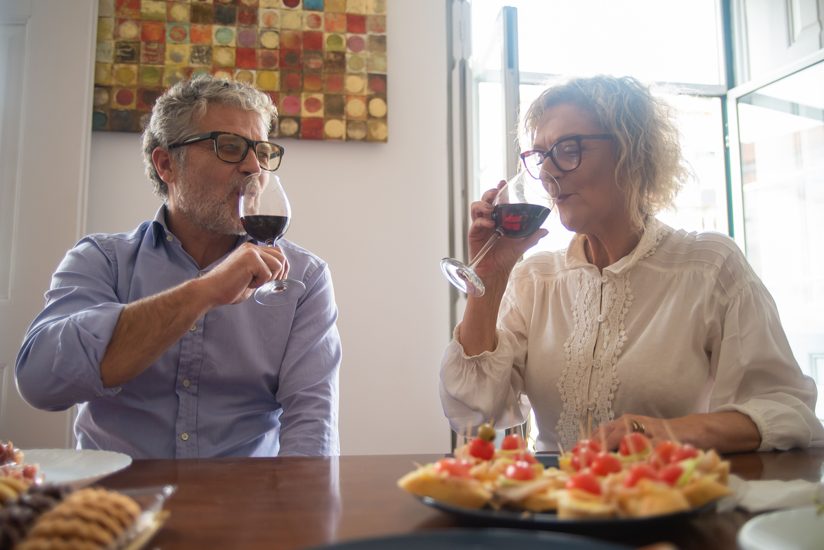 Casal sentado à mesa com aperitivos, aproximando do rosto duas taças de vinho tinto.