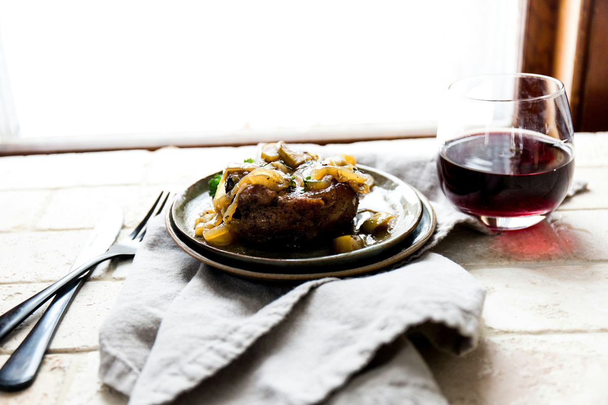 Carne suína coberta por cebolas e molho em prato escuro, com vinho tinto em taça sem haste ao lado.
