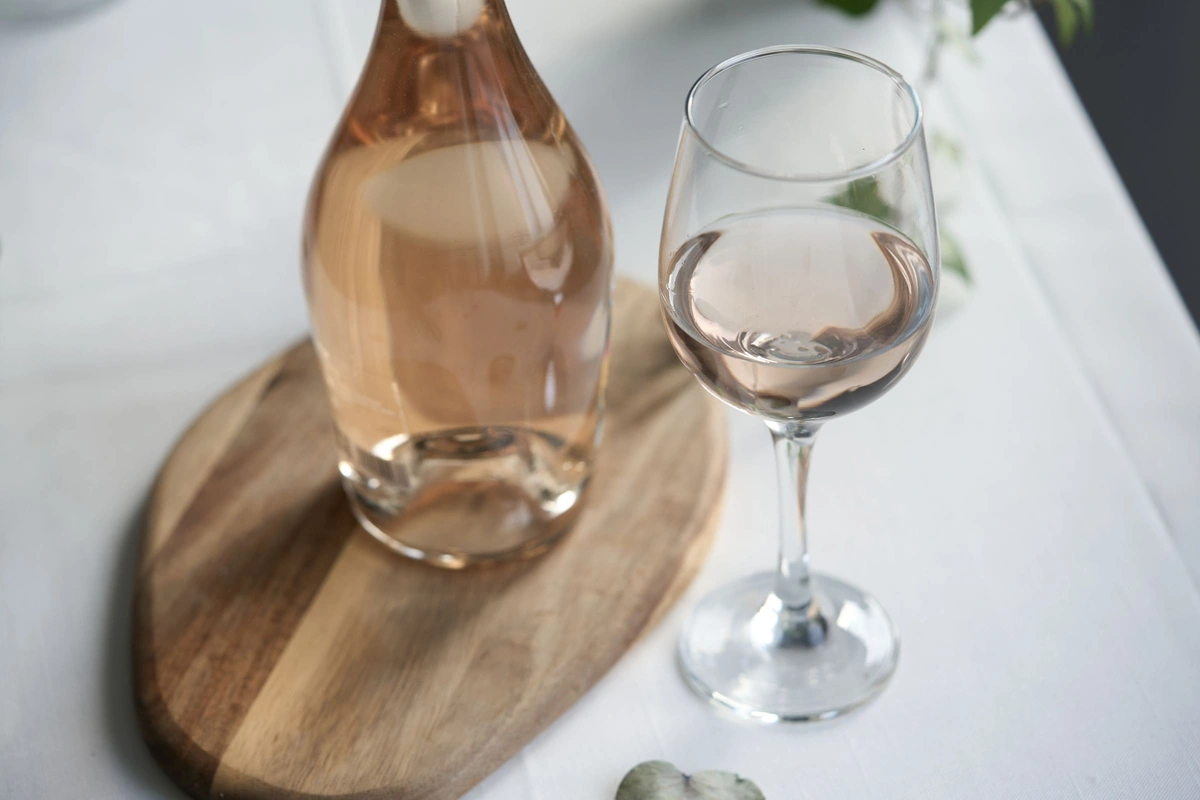 Taça de rosé claro em mesa clara, com garrafa sem rótulo visível ao fundo.