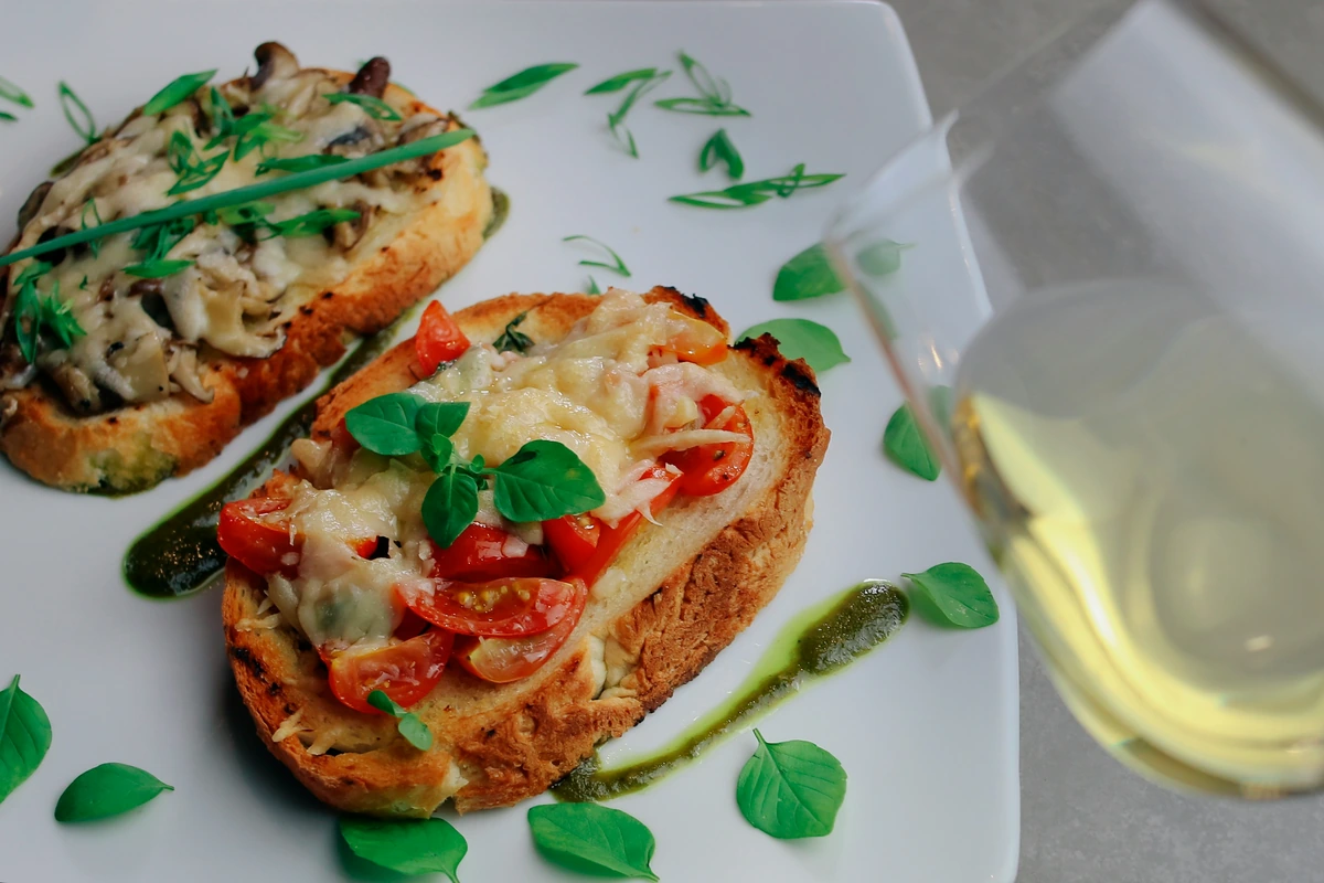 Bruschettas com tomate, queijo e cogumelos, ao lado de uma taça de branco.
