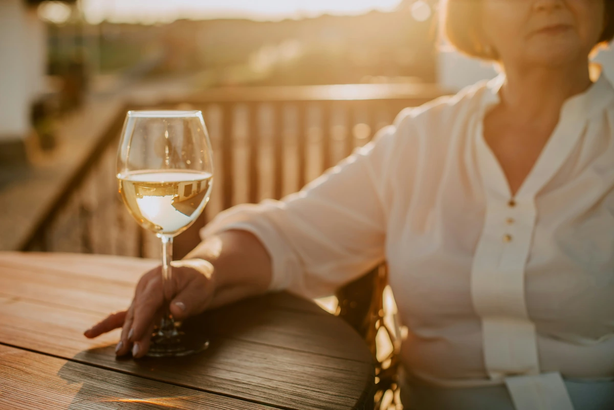 Mulher adulta de blusa branca segura uma taça de vinho branco claro em varanda de madeira, sob luz natural de fim de tarde.
