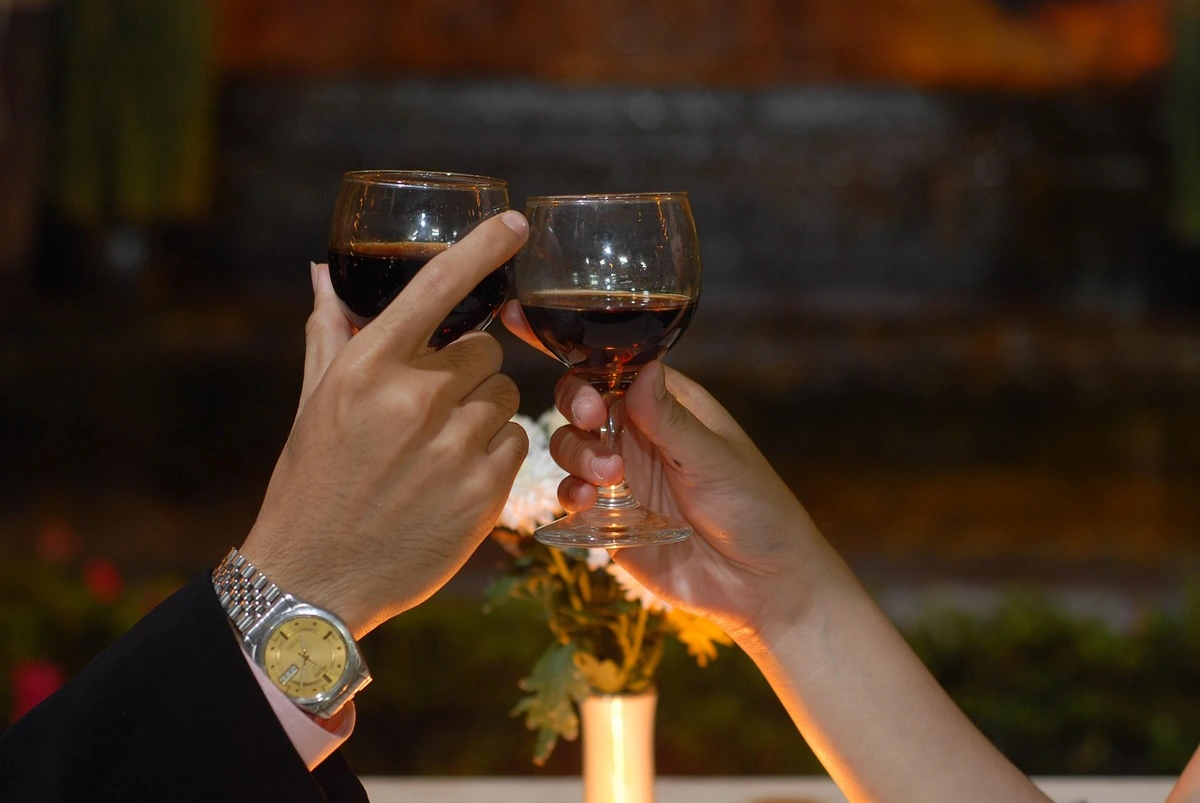 Duas mãos aproximando taças de vinho tinto com arranjo de flores ao fundo.