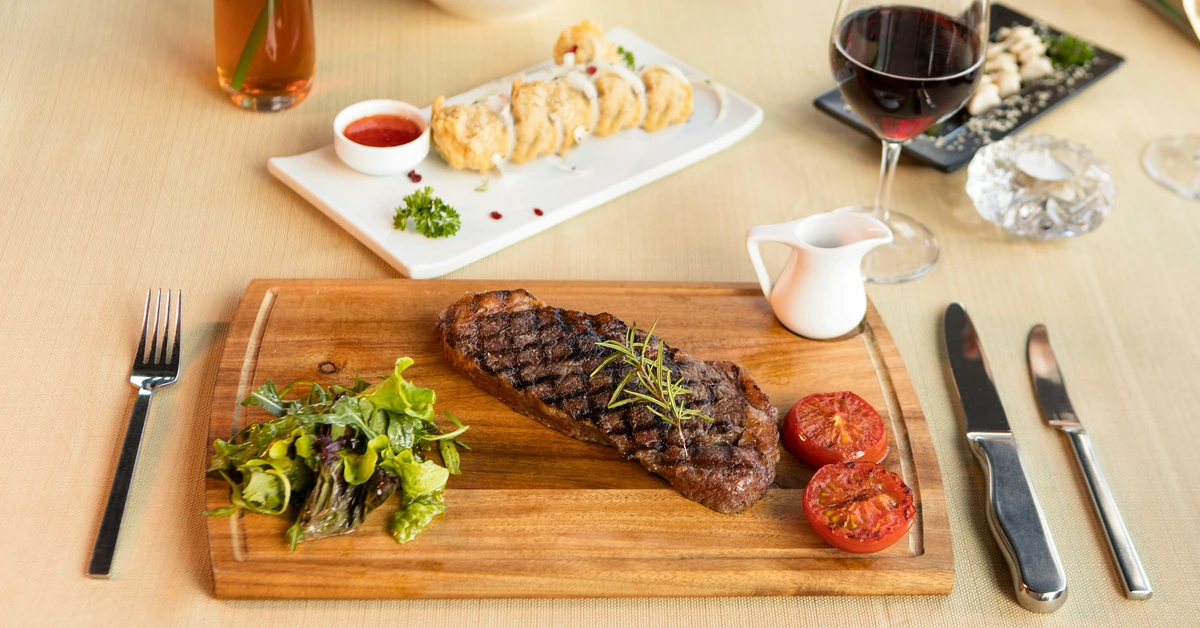 Bife grelhado sobre tábua com folhas e tomates; taça de vinho tinto à direita.