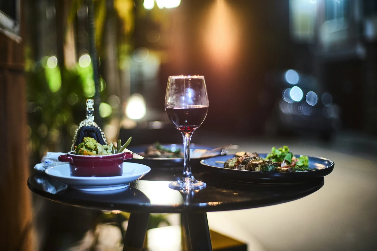 Taça de vinho tinto com pratos sobre uma mesa externa em luz quente.