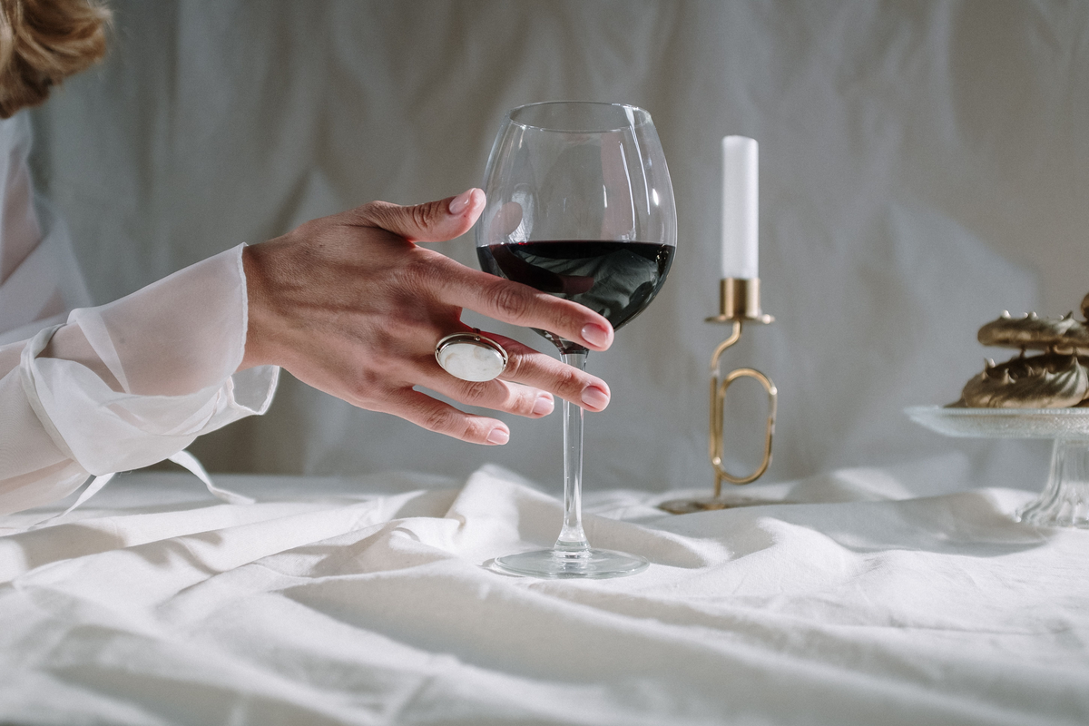 Mão junto a uma taça de vinho tinto sobre tecido branco, com castiçal e doces ao lado.