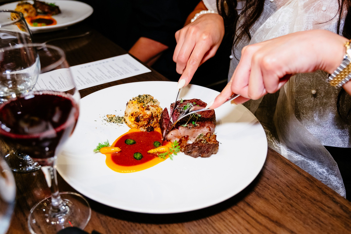 Pessoa corta carne com molho e legumes; taça de vinho tinto à esquerda.