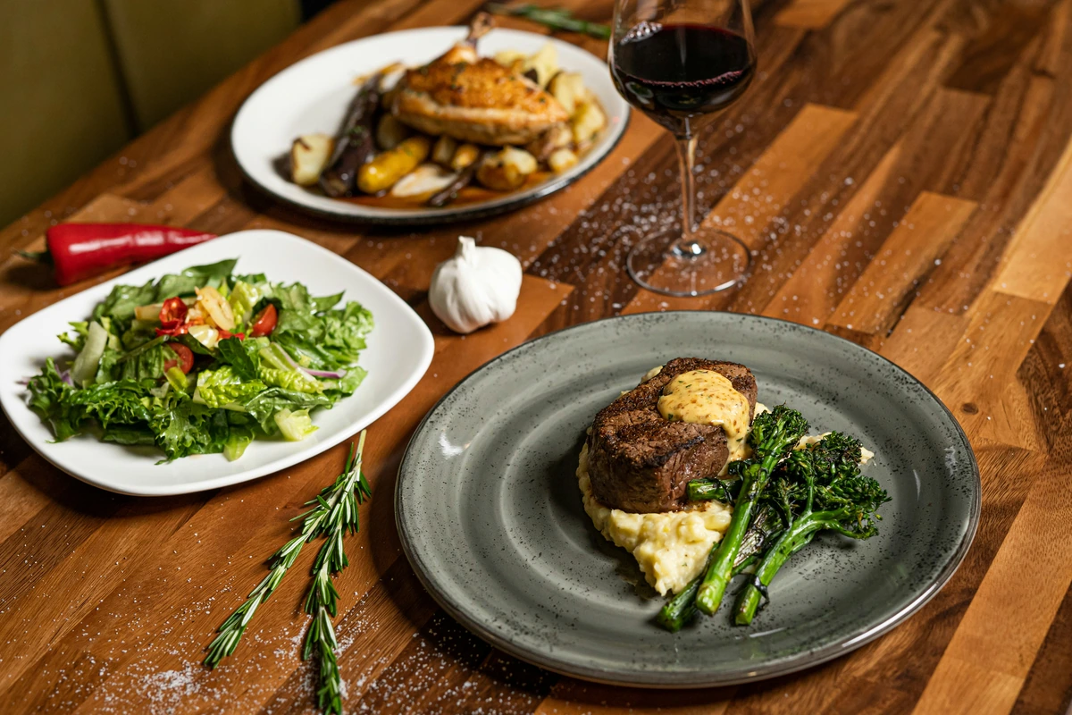 Carne com cobertura cremosa, purê e vegetais acompanhada de taça de vinho tinto.
