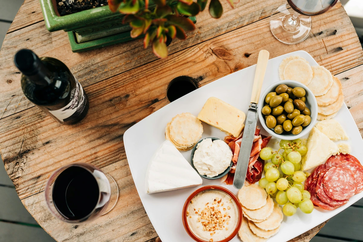 Queijos, charcutaria, uvas e azeitonas em travessa com uma taça de vinho tinto à esquerda.