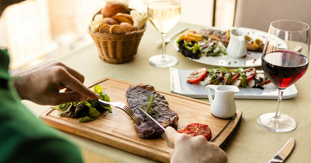 Steak com alecrim sendo cortado à mesa, com taça de tinto e acompanhamentos
